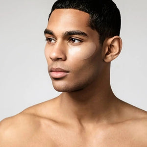 A young man with short dark hair and smooth skin looks to the side, wearing JAXON LANE - BRO MASK HYDROGEL COOLING EYE GELS 6 PACK by JAXON LANE to target dark circles. He is shirtless against a plain light background.