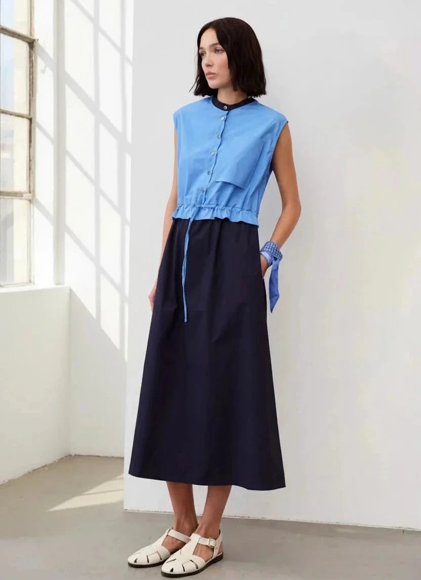 A model wears a sleeveless dress with a blue button-down bodice and a gathered waist above a navy midi skirt, paired with white closed-toe sandals.