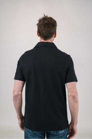 A man with light brown hair and a short beard wears the EDWARD ARMAH Pima Cotton Tailored Polo Shirt in dark blue and jeans, standing against a plain light background with a relaxed expression and one hand in his pocket.