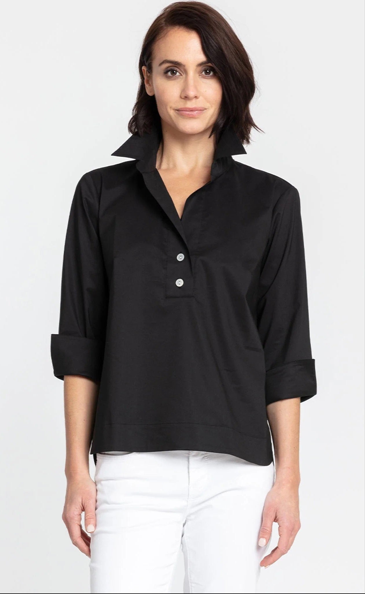 A person with short, dark hair is standing against a plain background, wearing the HINSON WU - AILEEN 3/4 SLEEVE TOP in black with a shirt collar and the sleeves rolled up, paired with white pants. The top features three buttons at the neckline. The individual is looking directly at the camera and smiling slightly.