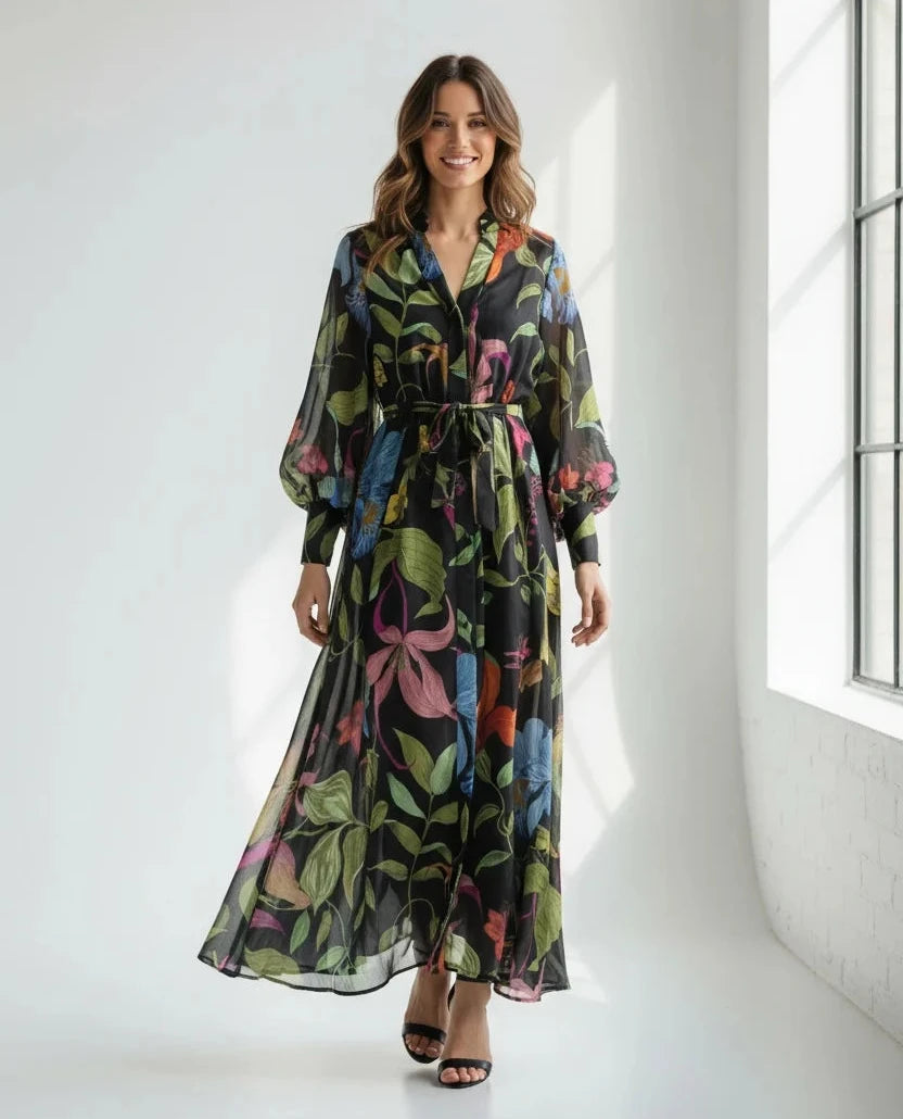 A woman wearing a black, long-sleeved, floor-length dress with a vibrant, multicolored floral and leaf print and a tie waist.