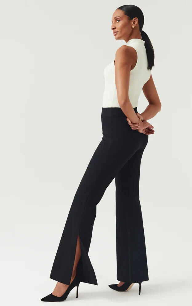 A woman stands posed against a light background, wearing the SPANX - PERFECT PANT SPLIT HEM, featuring black high-waisted pants crafted from smoothing premium ponte fabric with slit flares. Paired with a white sleeveless top and black high heels, her hair is pulled back as she looks to the side, hands clasped gracefully behind her back.