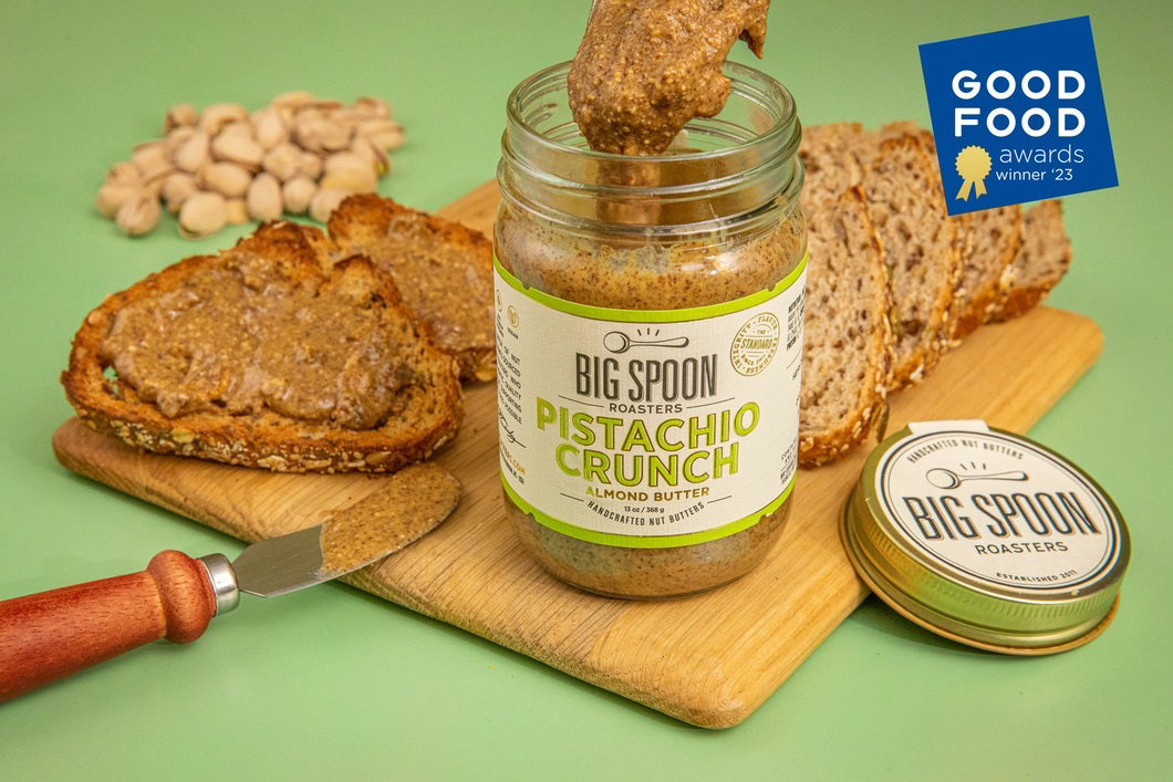 A jar of Big Spoon Roasters Pistachio Crunch almond butter sits open on a wooden board with a knife, toast, whole pistachios, and a lid nearby. A "Good Food Awards winner '23" badge is in the top right corner.