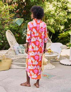 A person with curly hair stands outdoors, wearing the FRANK & EILEEN Rory Maxi Shirtdress in Classic Italian Linen—Orange and Pink Floral—embodying California chic amid lush greenery and white outdoor furniture, holding a woven straw bag.
