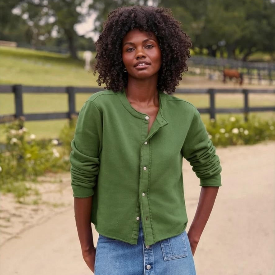 A woman stands outdoors on a dirt path, wearing the FRANK & EILEEN JACKIE Sweatshirt Cardigan in Triple Fleece Nori from the California Heritage Collection—a green button-up sweater paired with a blue denim skirt. There is a wooden fence, green grass, and trees in the background.