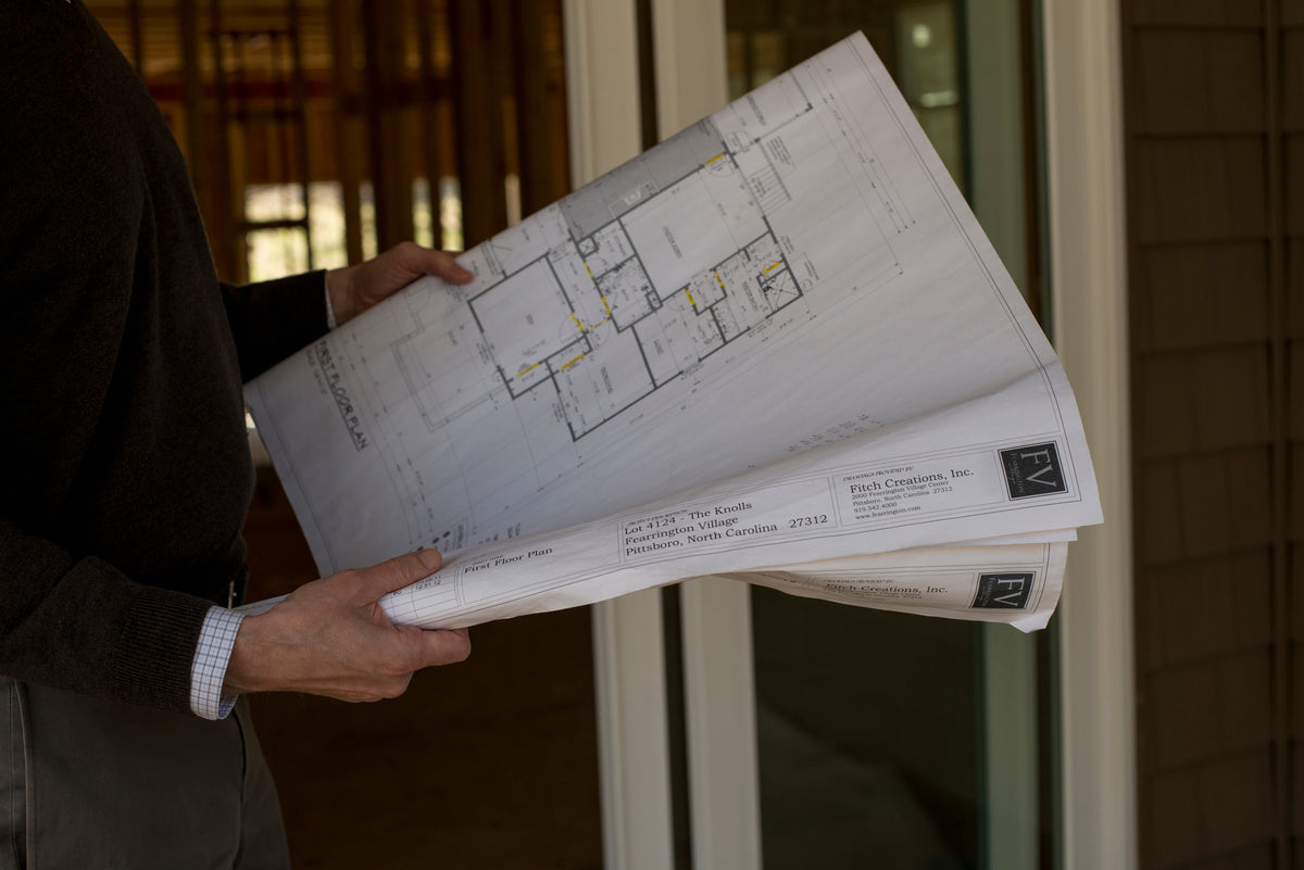 A person holding large architectural blueprints in both hands, standing inside a building under construction with wooden framing visible in the background.