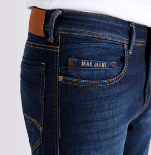 A person wearing MAC JEANS - BEN AUTHENTIC DENIM REGULAR FIT DARK VINTAGE WASH by MAC, paired with a white t-shirt and white sneakers, stands against a plain light gray background.