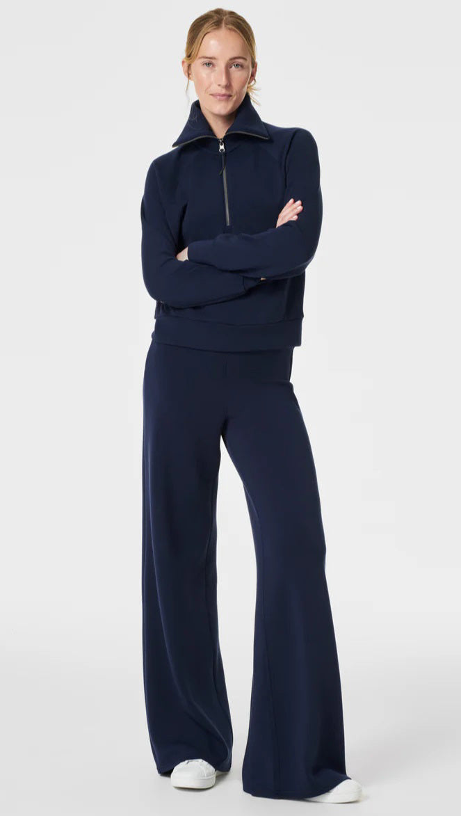 A person stands against a plain background wearing a matching two-piece outfit. The ultimate throw-on-and-go style consists of a long-sleeve, quarter-zip, high-collar pullover and SPANX AIRESSENTIALS WIDE LEG PANT in a deep blue color. They also wear white sneakers and have their hair pulled back.