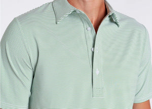 A man wears the CRIQUET Performance Sport Range Polo in Hobby Stripe - Gator, featuring light green and white stripes, a collar, and buttons, standing with one hand in his pocket against a plain background.
