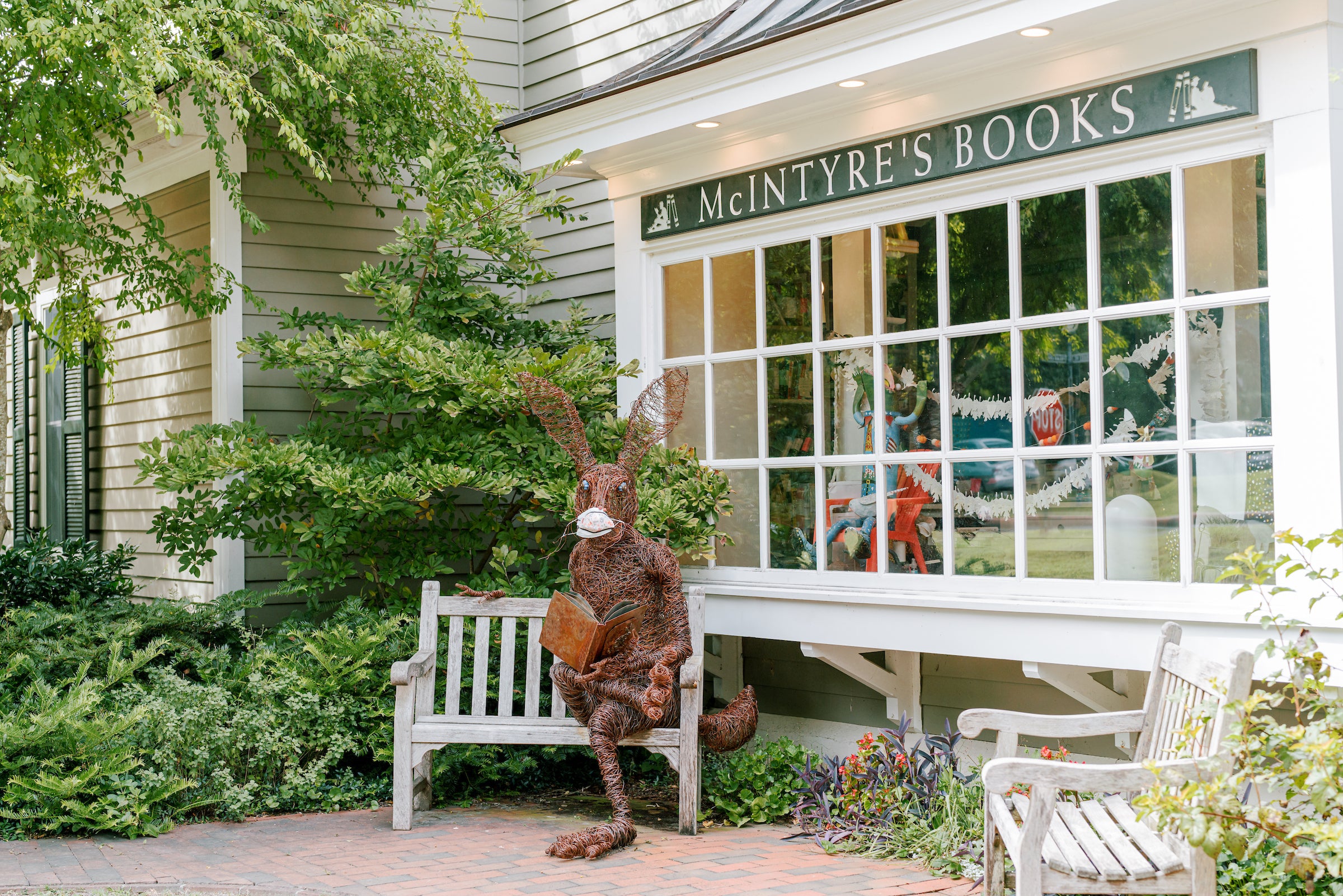 A large rabbit sculpture sits on a bench outside McIntyre’s Books, holding an open book. The store has big windows, and greenery surrounds the entrance. Another bench and colorful flowers are nearby.