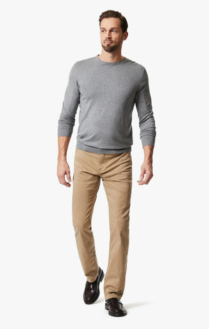 A man wearing a grey long-sleeve shirt, black dress shoes, and 34 HERITAGE - CHARISMA RELAXED STRAIGHT PANTS IN KHAKI TWILL stands with one hand in his pocket against a plain white background.