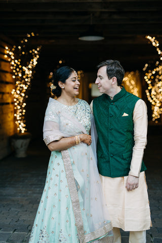 A smiling couple in traditional Indian attire stand together under string lights; the woman wears a pale blue and gold sari, while the man wears a cream kurta with a green vest. They look at each other, appearing joyful.