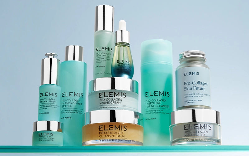 A collection of ELEMIS skincare products, including jars, bottles, and pumps in shades of blue, green, and silver, arranged on a reflective surface against a light blue background.