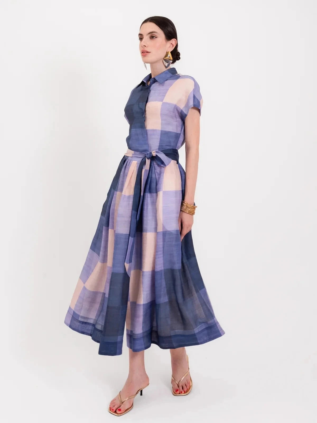 A model wears a short-sleeved, belted midi shirt dress featuring a large blue and beige plaid pattern.
