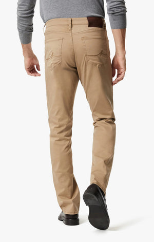 A man wearing a grey long-sleeve shirt, black dress shoes, and 34 HERITAGE - CHARISMA RELAXED STRAIGHT PANTS IN KHAKI TWILL stands with one hand in his pocket against a plain white background.