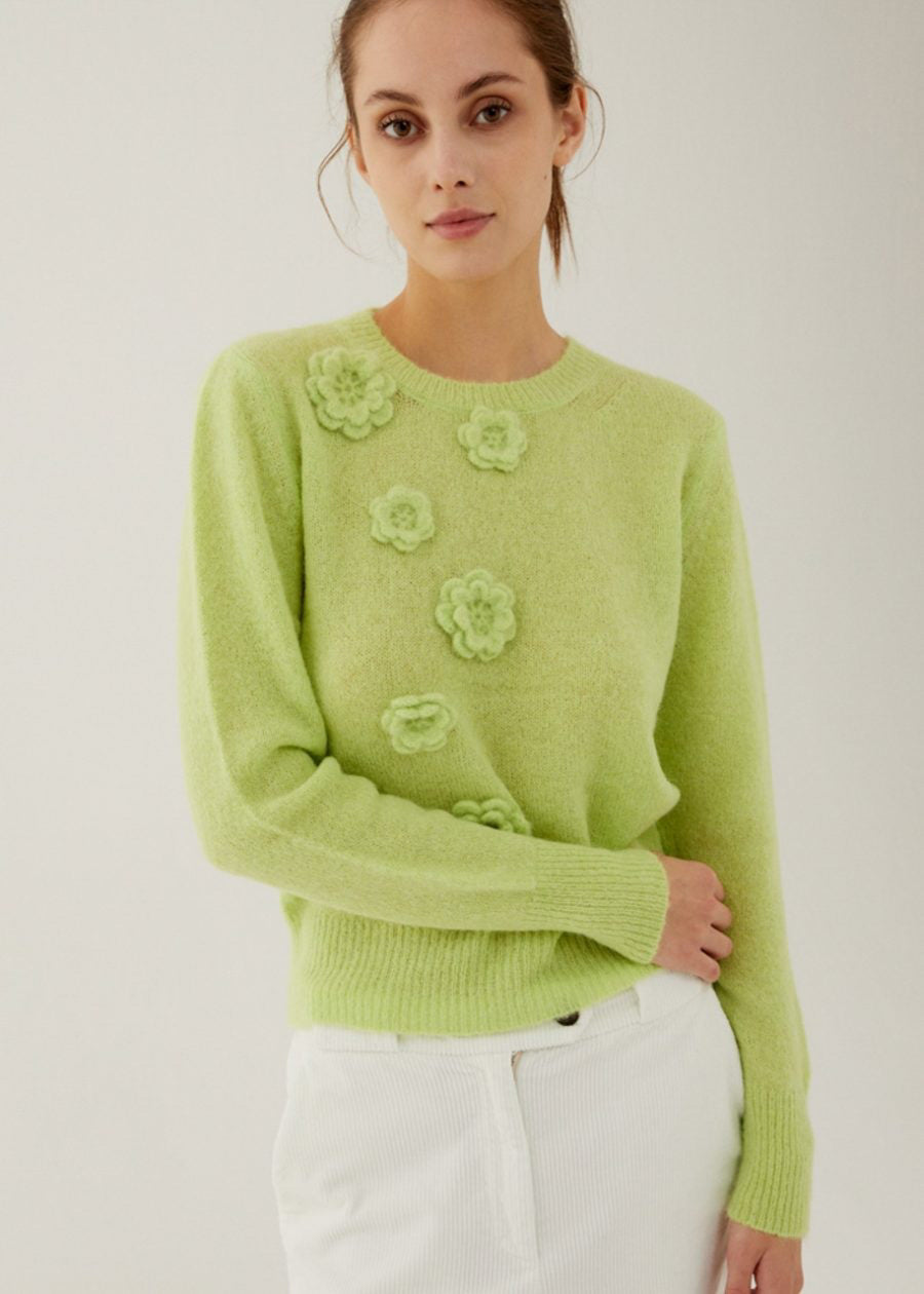 A woman wearing a light green sweater with 3D floral appliqués on the front and a white skirt stands against a plain light background, looking at the camera with a neutral expression.
