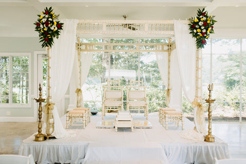 A beautifully decorated mandap with white drapes, floral arrangements, golden chairs, and traditional brass lamps set up indoors for a Hindu wedding ceremony, with large windows showing greenery outside.