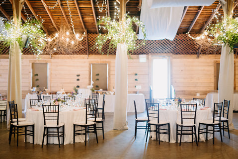 A rustic wedding reception hall with round tables covered in white cloths, black chairs, and greenery decorations. String lights and chandeliers hang from a wooden ceiling, creating a warm, festive atmosphere.