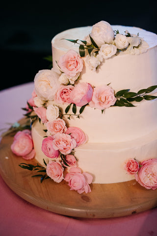 A two-tier white cake decorated with cascading pink and white roses and greenery, displayed on a wooden board with a pink tablecloth underneath.
