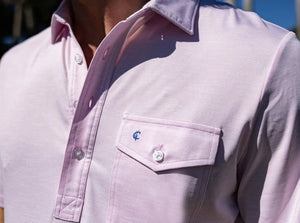 A man with a beard is wearing the CRIQUET Performance Sport Heather Players Shirt in Pink Panther and pale blue pants, standing against a plain white background.