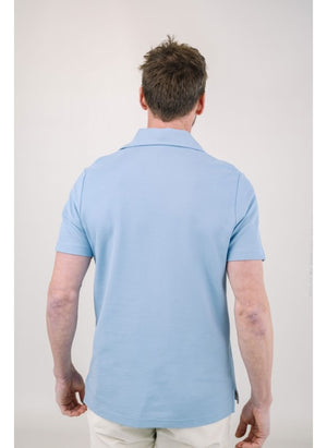 A man with light brown hair and a short beard wears the EDWARD ARMAH Pima Cotton Tailored Polo Shirt in dark blue and jeans, standing against a plain light background with a relaxed expression and one hand in his pocket.
