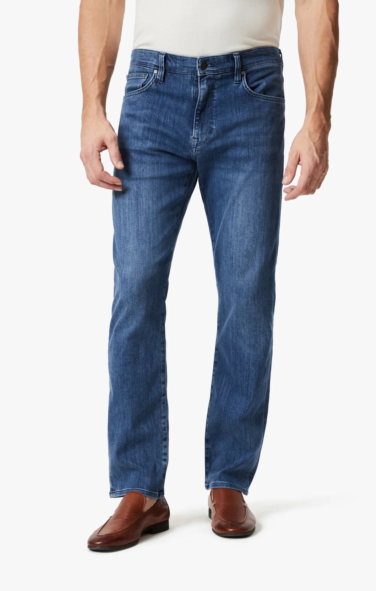 A man wears 34 HERITAGE CHARISMA Relaxed Straight Leg Denim in Mid Urban Wash, a white shirt, and brown leather shoes, standing against a plain white background.