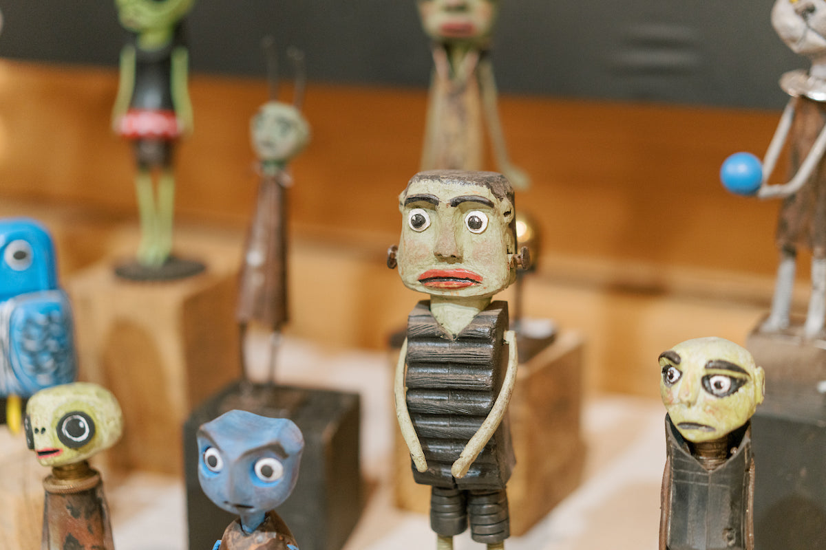 A group of quirky, colorful wooden figurines with exaggerated facial expressions and features are displayed together, each with distinct painted faces and unique shapes, standing on wooden blocks.