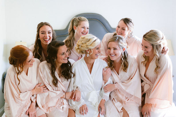 Seven women in light pink robes sit and smile around a woman in a white robe, sharing a joyful moment together on a bed, likely getting ready for a wedding.
