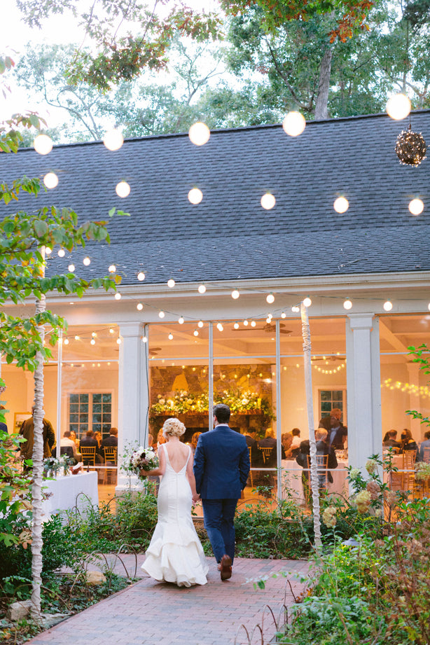 A bride and groom walk hand in hand down a garden path toward a warmly lit building, decorated with hanging string lights, as wedding guests are seen inside through large windows.