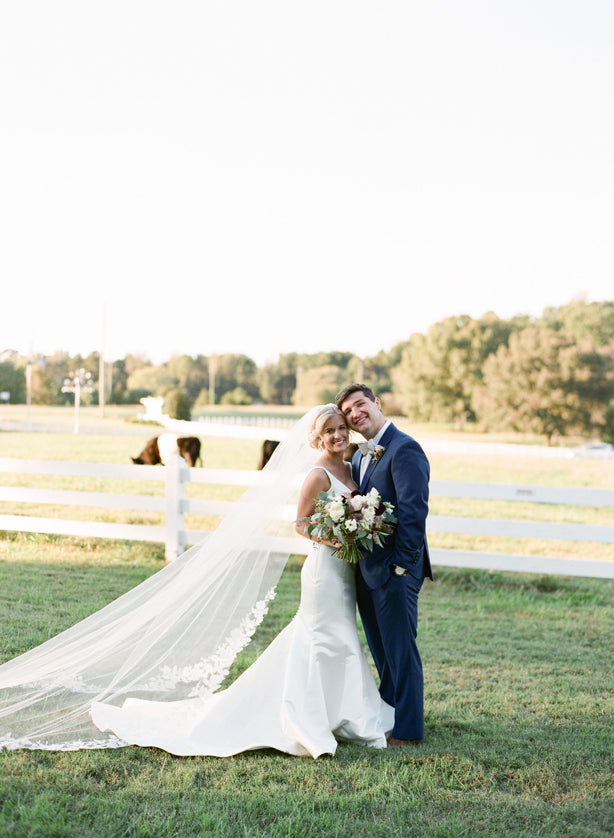 A bride and groom stand smiling together outdoors on grass near a white fence, with the bride’s long veil flowing behind her. The bride holds a bouquet and cows graze in the background under a bright sky.