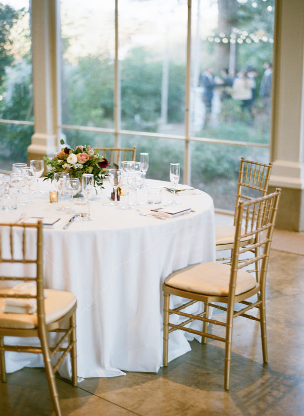 A round table with a white tablecloth is set with glassware, silverware, and a floral centerpiece. Four gold chairs surround the table. Large windows in the background reveal a garden scene outside.