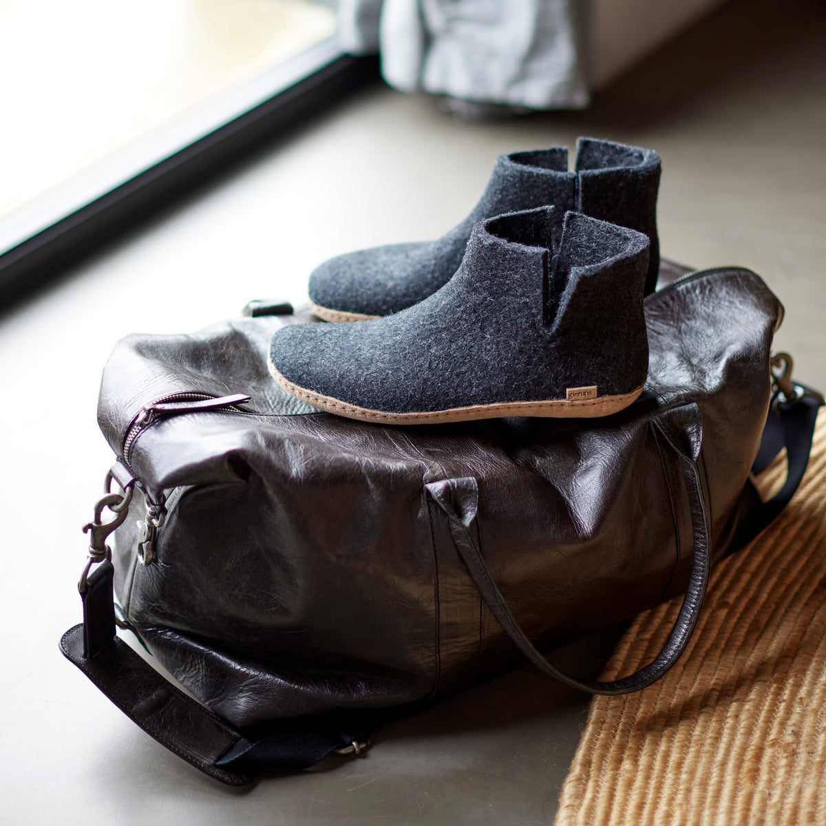 A pair of dark gray felt ankle boots rests on top of a black leather duffel bag placed on a floor next to a window and a woven rug.