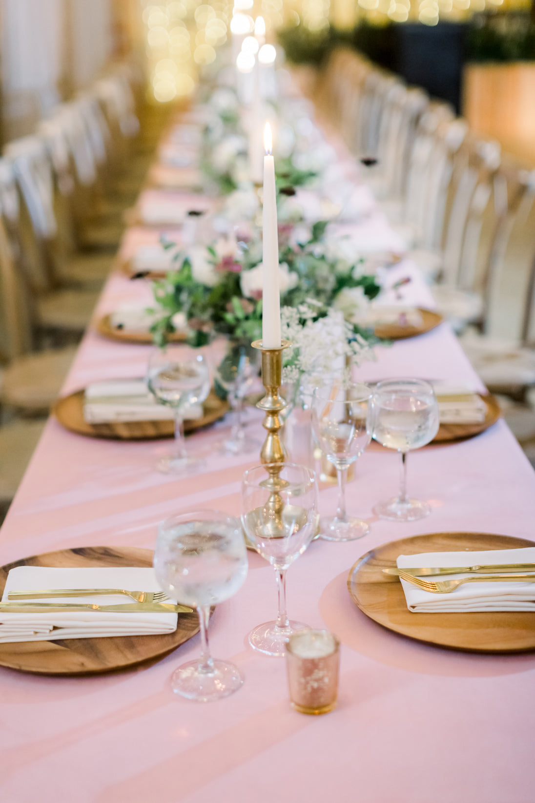 A long banquet table with a pale pink tablecloth, set with wooden plates, gold cutlery, wine glasses, and tall white candles in gold holders, decorated with floral centerpieces and soft, romantic lighting.