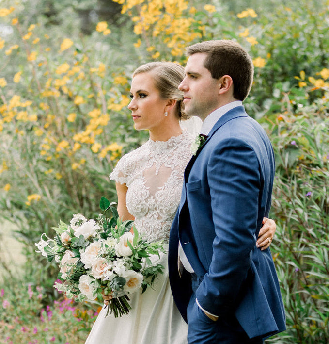 A bride in a white dress holding a bouquet stands beside a groom in a blue suit. They embrace outdoors, surrounded by greenery and yellow flowers, looking thoughtfully into the distance.