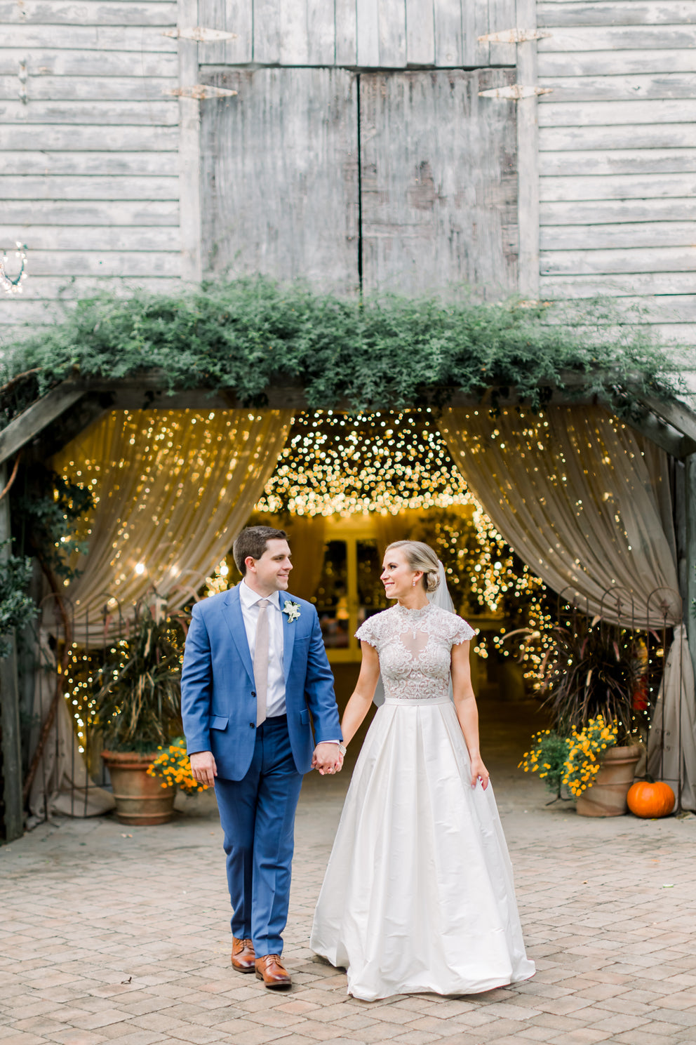 A bride and groom hold hands and smile at each other while walking outside a rustic barn decorated with string lights, greenery, and yellow flowers. The bride wears a white gown; the groom wears a blue suit.