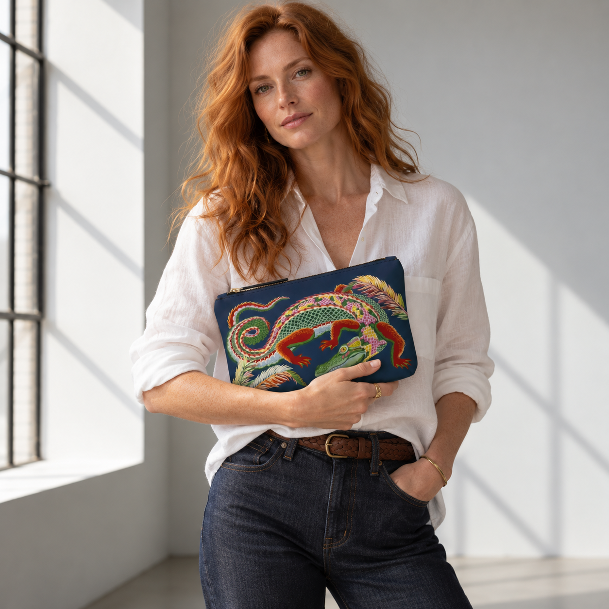 A woman in a white button-down shirt and jeans holds a navy blue clutch featuring a colorful embroidered lizard.