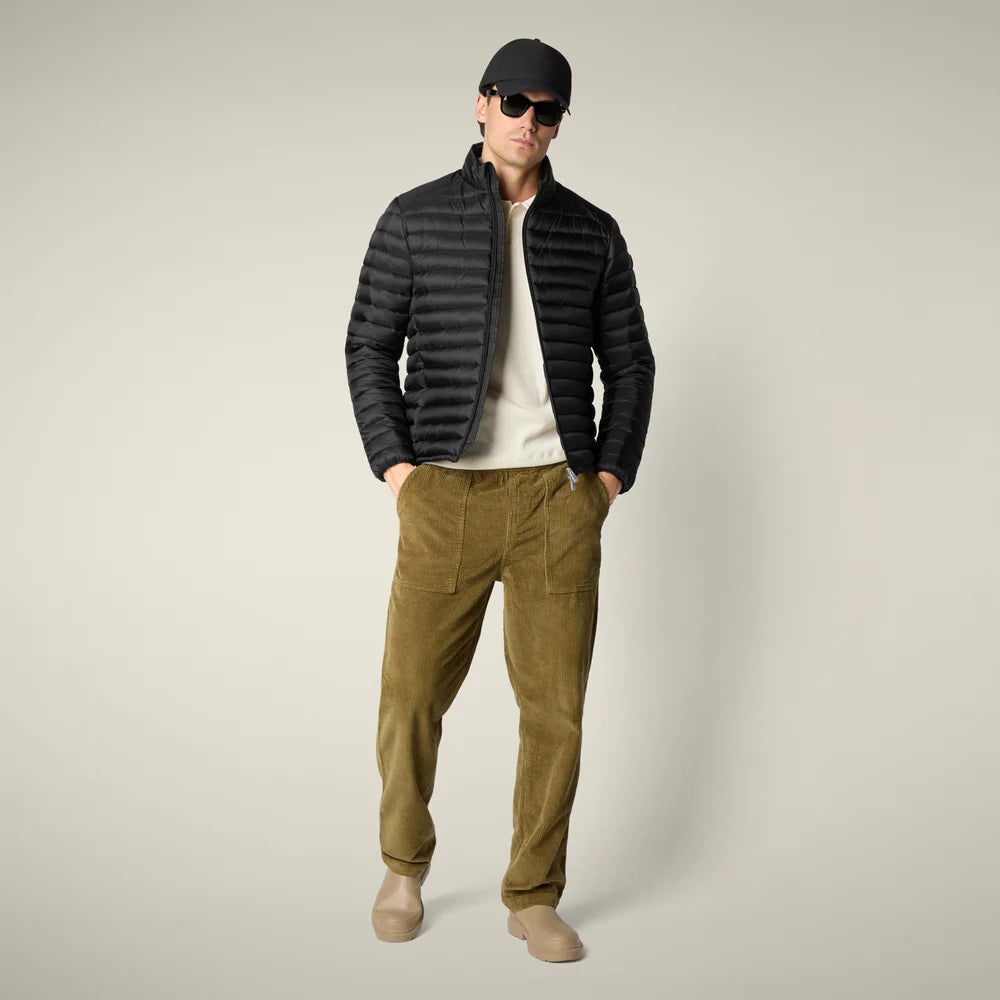 A person wearing a black puffer jacket over a white shirt, tan corduroy pants, taupe shoes, a black cap, and black sunglasses stands against a plain light background.