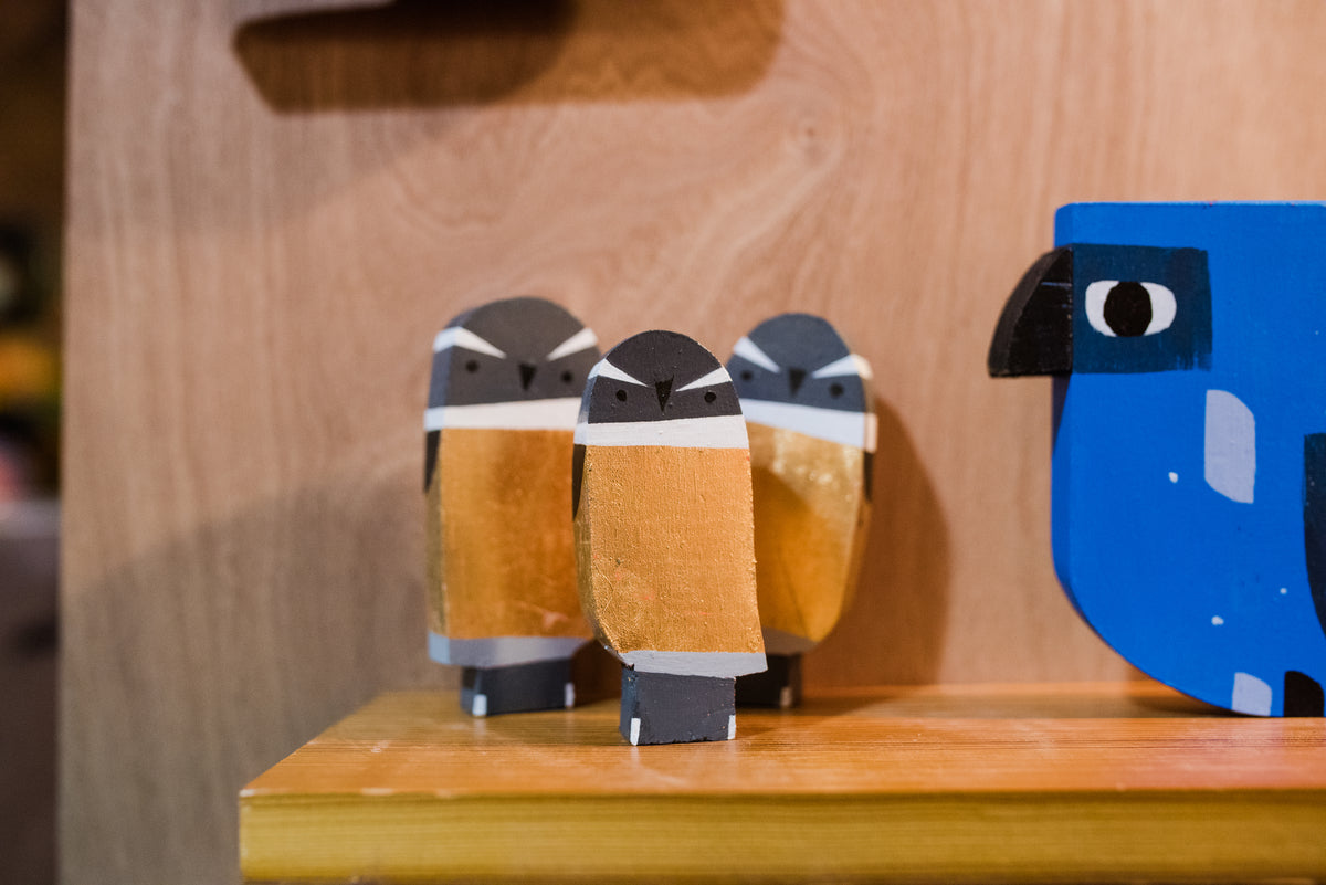 Three small, hand-painted wooden bird figurines with brown, white, and black coloring stand on a wooden shelf beside a larger, blue bird-shaped object.