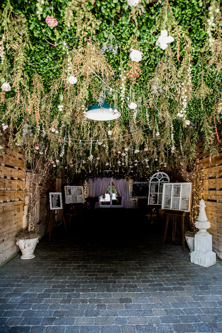 A rustic hallway with wooden walls and brick flooring, decorated with hanging greenery, flowers, and string lights, leading to a softly lit room with vintage frames and white drapes at the far end.