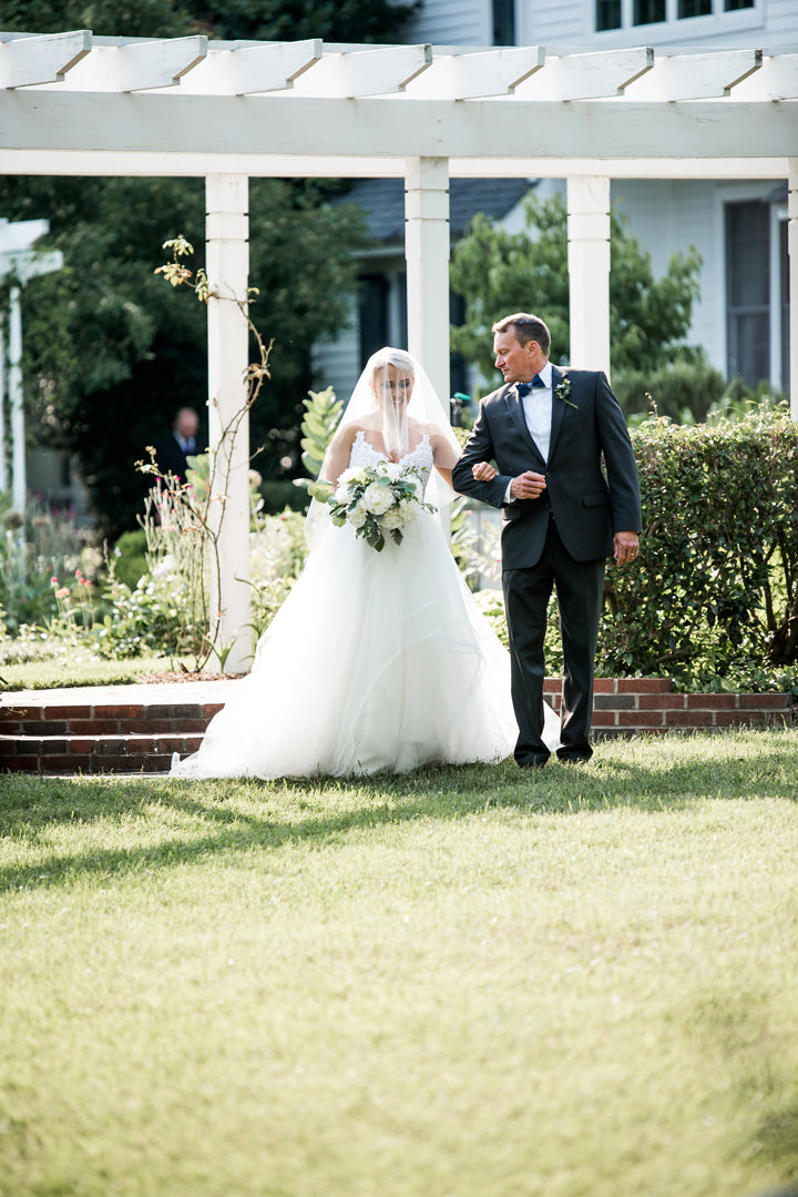 A bride in a white gown and veil holds a bouquet while walking on grass, arm-in-arm with a man in a suit. They are outdoors near a white pergola and greenery, with sunlight shining.