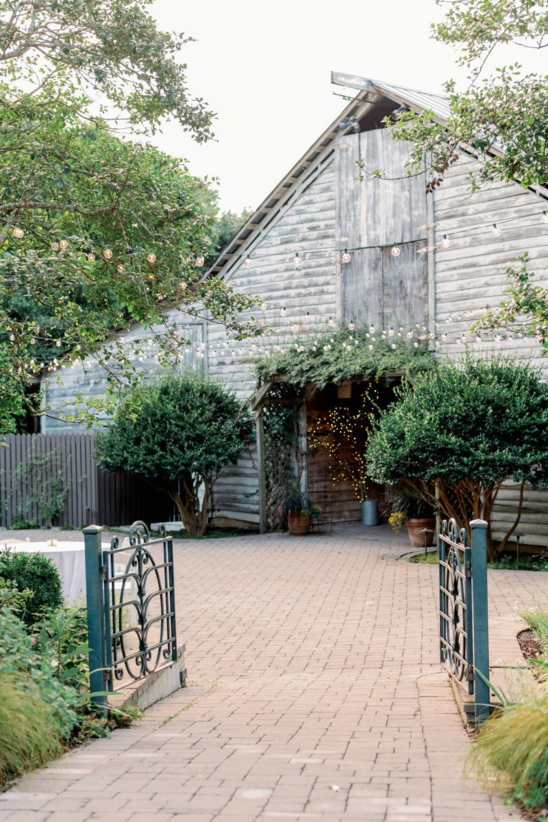 A rustic wooden barn with a sloped roof is decorated with string lights and surrounded by green trees and bushes. A brick walkway leads to the entrance, creating a welcoming, festive atmosphere.