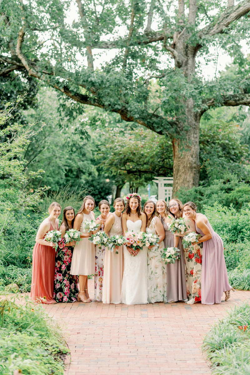 A bride in a white dress stands outdoors on a brick path, surrounded by ten smiling bridesmaids in colorful, floral, and pastel dresses. They each hold bouquets, with green trees and lush garden scenery in the background.