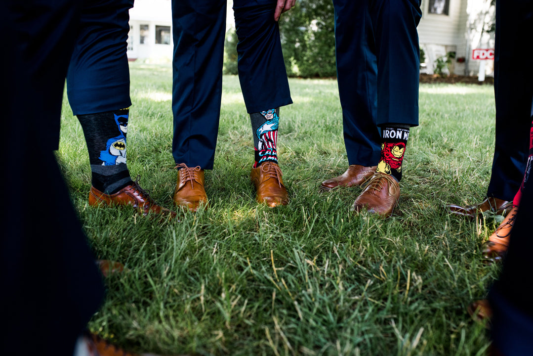 Four people in dress shoes and dark blue suits stand on grass, showing off superhero-themed socks featuring Batman, Captain America, and Iron Man. The focus is on their socks and lower legs.