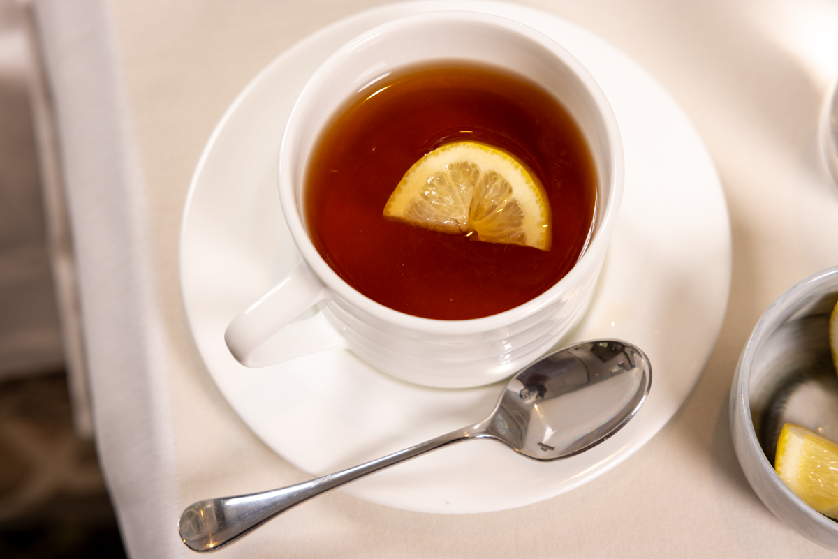 A white cup of tea with a slice of lemon inside, placed on a white saucer with a metal spoon beside it, all set on a light-colored tablecloth.