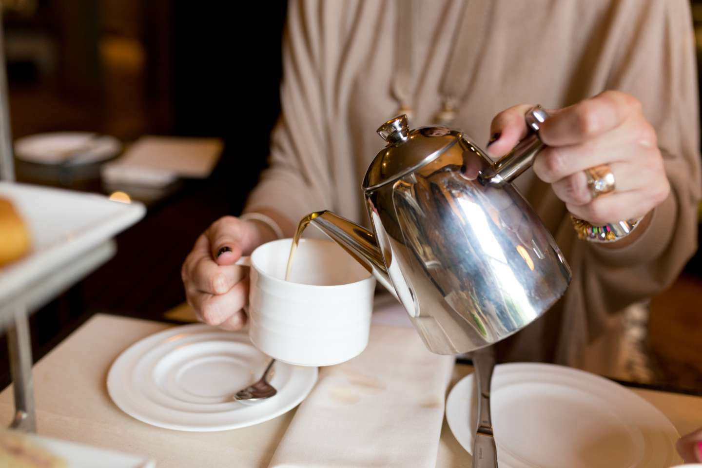 A person pours tea from a shiny silver teapot into a white teacup on a saucer. The table is set with plates, a spoon, and a napkin. The person is wearing a beige top and a ring on their right hand.