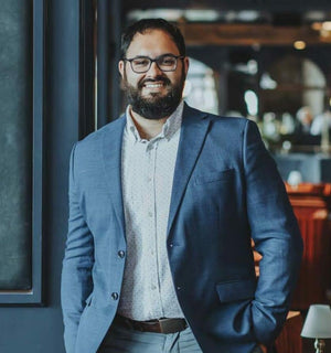 A bearded man with glasses, dressed in a blue suit jacket and light shirt, stands smiling in a warmly lit modern restaurant—Jonathan Ross, MS, Master Sommelier and guide for the Legend Wine Dinner with FEARRINGTON VILLAGE.