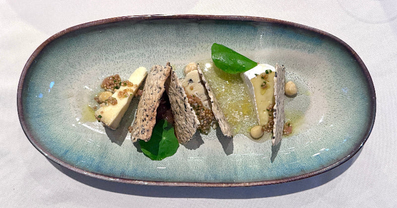 A rectangular ceramic plate with slices of soft cheese, seeded crackers, green leaves, and dollops of yellow sauce, arranged neatly for a stylish presentation.