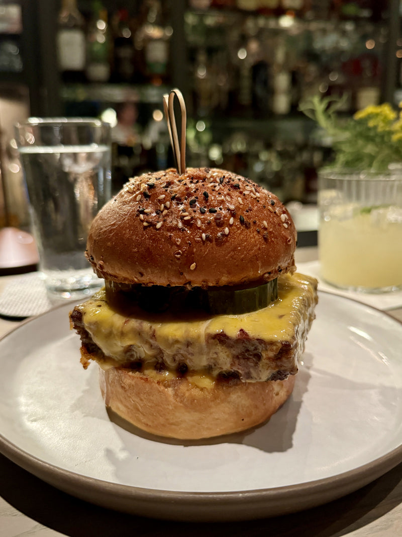 A close-up of a cheeseburger with a seeded bun, melted cheese, and a pickle slice on top, served on a white plate with a glass of water and another drink in the background.