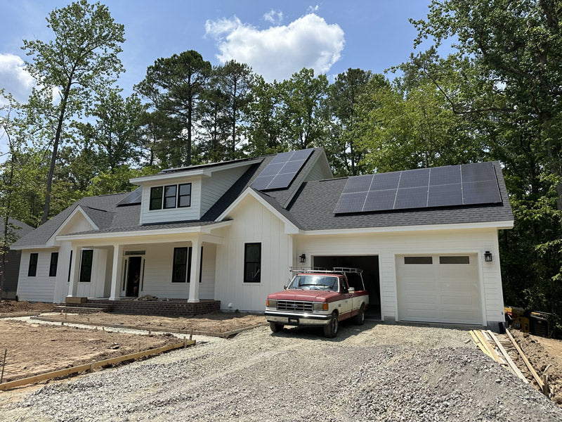 A modern white house with black roof shingles and solar panels on the roof, surrounded by trees. A red and white pickup truck is parked in the gravel driveway next to the garage.