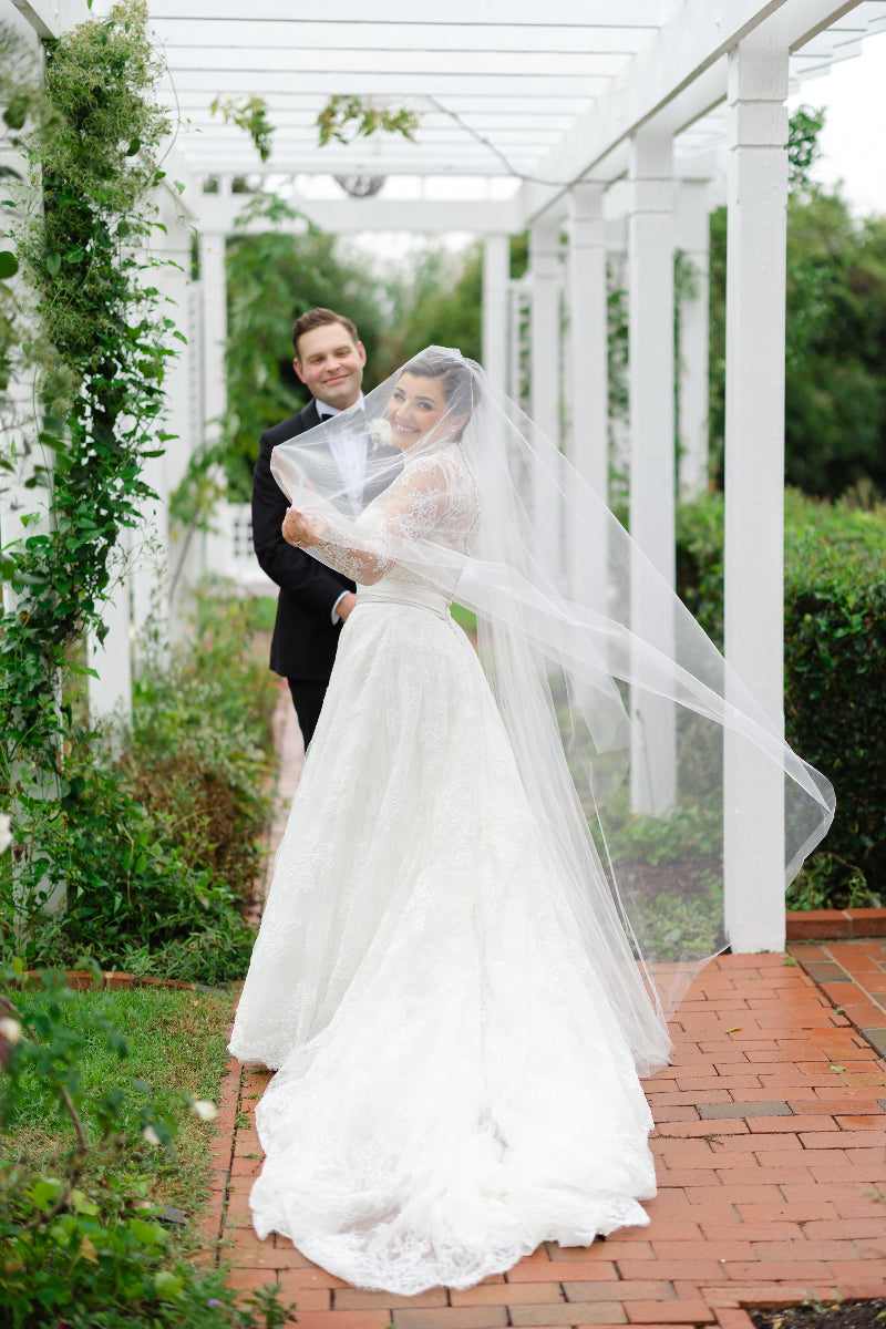 A bride in a white lace gown and long veil smiles back at the camera while holding hands with a groom in a black suit, standing under a white pergola surrounded by greenery.